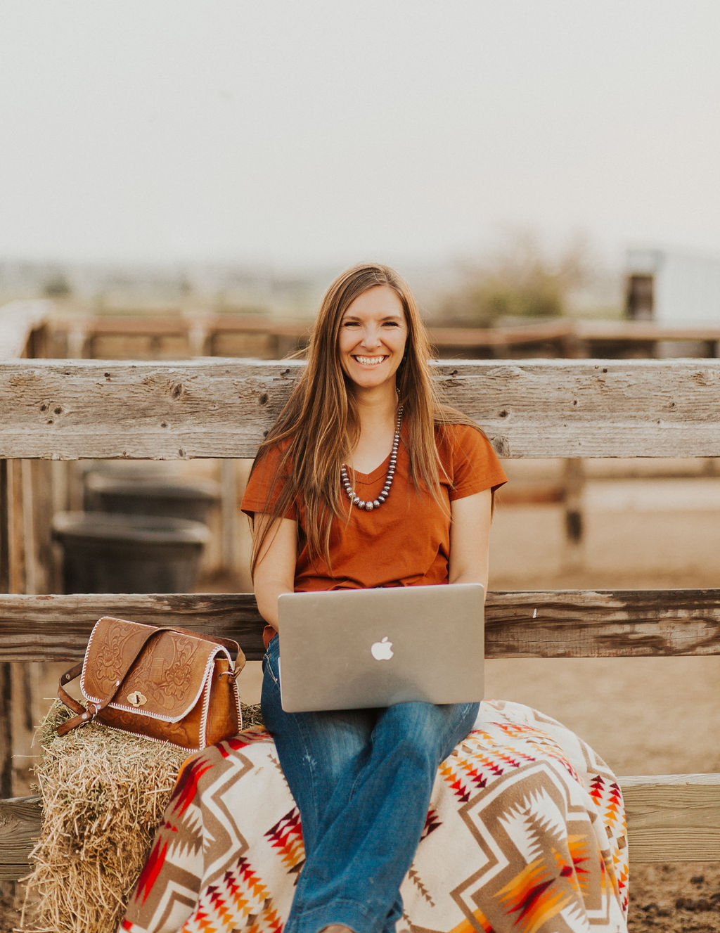 Woman with long dark blonde hair sitting on a hay bale and pendleton blanket with a laptop